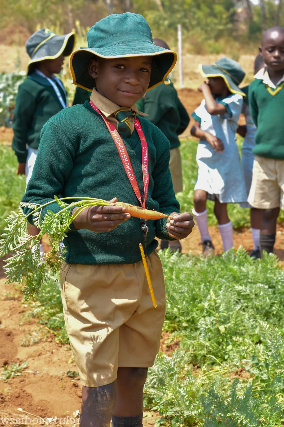A Nyamhuka learner in green uniform and bush hat holds up a carrot in the school garden